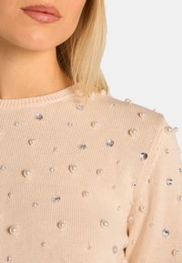 Beige gestrickter Pullover mit gleichmäßig verteilten Perl- und Strassverzierungen auf dem Stoff. Klassischer Rundhalsausschnitt.