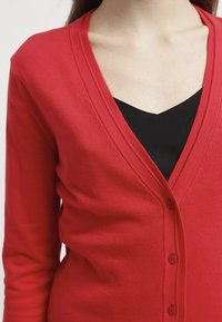 Röd kofta med v-ringning och långa ärmar, med en mjuk stickad textur och en rad av sex knappar fram.