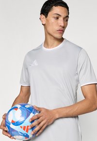 Ανοιχτό γκρι αθλητική μπλούζα με λευκό περίγραμμα, που διαθέτει τρεις διαγώνιες ρίγες στο αριστερό στήθος, συνδυασμένη με μια μπλε και λευκή μπάλα ποδοσφαίρου με σχέδιο.