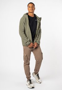 Groene fleece zip-up hoodie over een zwart t-shirt, gecombineerd met lichtbruine cargo shorts met zijzakken en beige sneakers met zwarte veters.