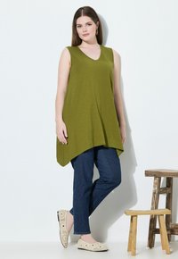 Tunica senza maniche verde oliva, scollatura a V, orlo asimmetrico, abbinata a jeans blu scuro e ballerine beige con fantasia. Sgabelli di legno sullo sfondo.
