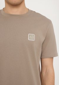 Beige tričko s krátkým rukávem vyrobené z měkké bavlny. Na hrudi má malé čtvercové logo s textem "A|X" v kontrastních barvách.
