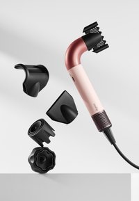 Dyson-hårfön med ett böjt rosa handtag och svarta accenter. Har flera avtagbara svarta munstycken på en ljusgrå yta.