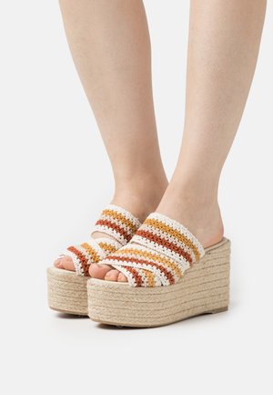 Pieds portant des sandales compensées à plateforme avec des semelles tressées beiges et des dessus au crochet rayés dans des tons blancs, orange et marron.