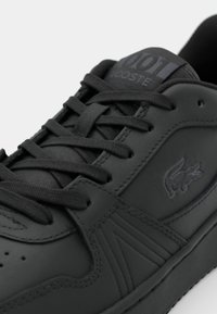 Sorte lær sneakers med en glat overflade, der har flade sorte snørebånd, prægede logo og minimalistiske designelementer på siden.