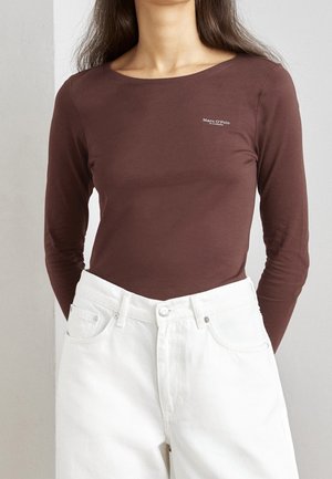 Langærmet brun bluse lavet af blødt stof med en rund halsudskæring og et lille logo på brystet, parret med hvide højtaljede jeans.