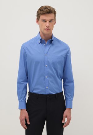 Young man wearing a blue button-up shirt with front pocket and black trousers, standing against a plain light background.