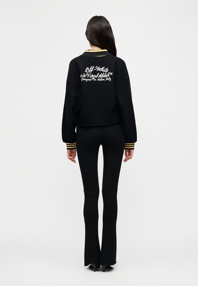 Μαύρο φούτερ με κολάρο και μανσέτες με κίτρινες ρίγες, στο πίσω μέρος υπάρχει λευκό κεντητό κείμενο. Συνοδεύεται από στενά μαύρα παντελόνια με φαρδύ τελείωμα που ολοκληρώνουν το σύνολο.