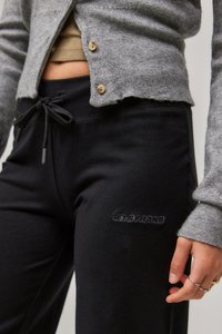 Schwarze Jogginghose aus weichem Stoff, mit elastischem Bund und Kordelzug sowie einem in Schwarz gestickten Logo. Graue Strickjacke sichtbar.