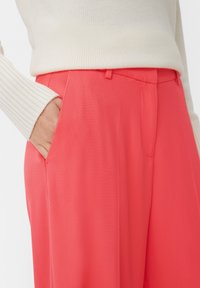 Pink, tailored trousers with a smooth texture, featuring side pockets and a subtle front crease. Styled with a cream ribbed sweater.