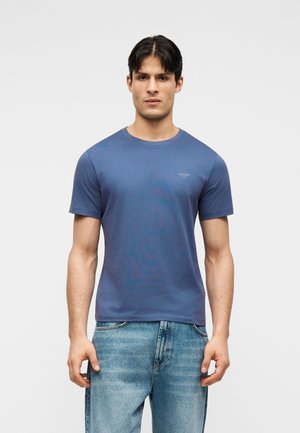 Moški, oblečen v preprosto modro majico s kratkimi rokavi in svetlo modre kavbojke, stoji ob enobarvnem svetlem ozadju.