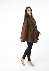 Capa marrón de estilo envolvente con acabado texturizado, con cinturón de lazo en la cintura; combinada con leggings negros y tacones transparentes.