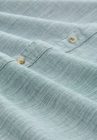 Tela de lino azul claro con un tejido de textura fina; presenta pequeños botones de color crema y sutiles líneas horizontales.