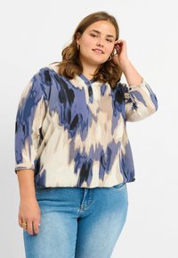 Blusa a maniche lunghe, dal taglio morbido, con un motivo astratto nei colori crema, blu e nero. Presenta un collo con bottoni e un tessuto morbido e leggero.