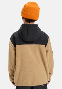 Giacca in fleece con una sezione inferiore beige e una sezione superiore nera. Presenta un cappuccio e un tessuto morbido e strutturato. Include un logo all'orlo.