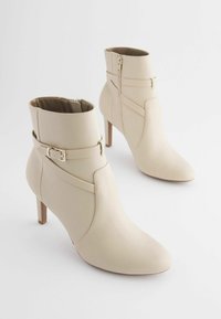 Stivaletti beige realizzati in pelle liscia, con punta affusolata, chiusura laterale con zip e una fascia decorativa con fibbia. Design con tacco a stiletto.