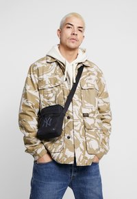 Giacca mimetica beige e bianca sopra un hoodie bianco, abbinata a jeans scuri. Borsa a tracolla nera con logo grigio. Capelli corti e biondi chiari.