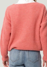 Sweter z długim rękawem, luźny, wykonany z teksturowanej koralowej przędzy, z szerokim dekoltem i ściągaczami przy rękawach, zestawiony z jasnoniebieskimi dżinsami.