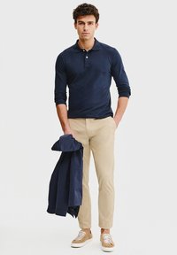 Granatowa koszulka polo z długim rękawem, beżowe chinosy, jasnobrązowe sneakersy. Trzyma granatową kurtkę, stojąc na neutralnym tle.