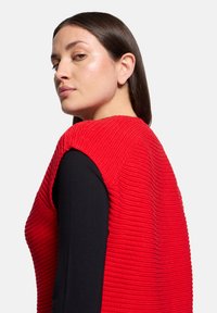 Czerwony, bezrękawnik o fakturowanej powierzchni z prążkowanym wzorem dzianiny, zestawiony z czarną koszulą z długim rękawem. Widok z boku podkreśla nowoczesną sylwetkę.