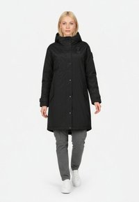 Sort lang jakke med hætte, der har lynlås og trykknaplukning, sidelommer og en glat tekstur, parret med grå bukser og hvide sko.