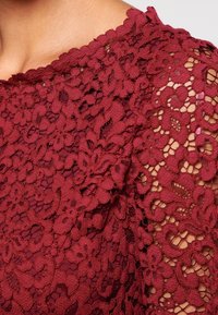 Burgundy pitsakanka, joka esittelee kukkakuvioita, halkioitu kaula-aukko ja läpikuultavat yksityiskohdat hihoissa luoden monimutkaisen tekstuurin.
