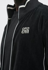 Casaco preto canelado com fecho éclair e logótipo branco "OPEN ERA" no peito, e gola às riscas, usado por cima de camisa preta.