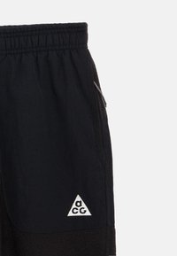 Melnas bikses ar elastīgu jostasvietu, sānu kabatu ar rāvējslēdzēju un trīsstūra baltu logotipu uz apakšējās kājas, apvienojot gludu un teksturētu audumu.