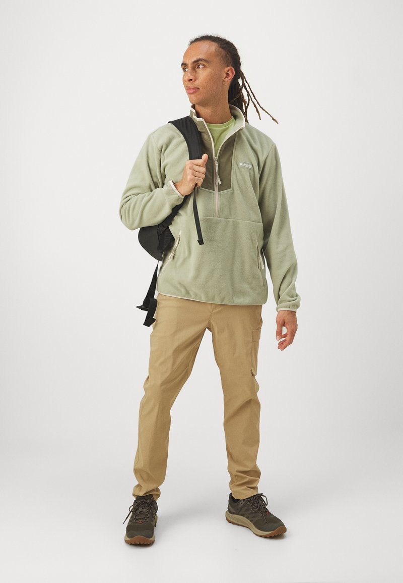 Fleecetrekker in lichtgroen met een halve rits, zijzakken; gecombineerd met beige cargobroeken en donkere wandelschoenen. Minimalistisch ontwerp.