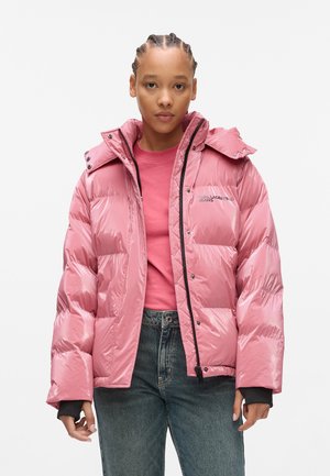 Veste rose matelassée avec une finition brillante, dotée d'une capuche, d'une fermeture éclair et d'accents noirs. Portée par-dessus une shirt rose et un jean bleu.