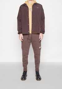 Hnědý větrový kabát se zapínáním na tlakové knoflíky, přes žlutou mikinu. Hnědé tepláky s bočními kapsami a bílými bočními pruhy. Černé boty na šněrování.