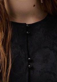 Svart, texturerad klädsel med rundad halsringning och tre dekorativa svarta knappar längst fram; närbild av tygdetaljer.