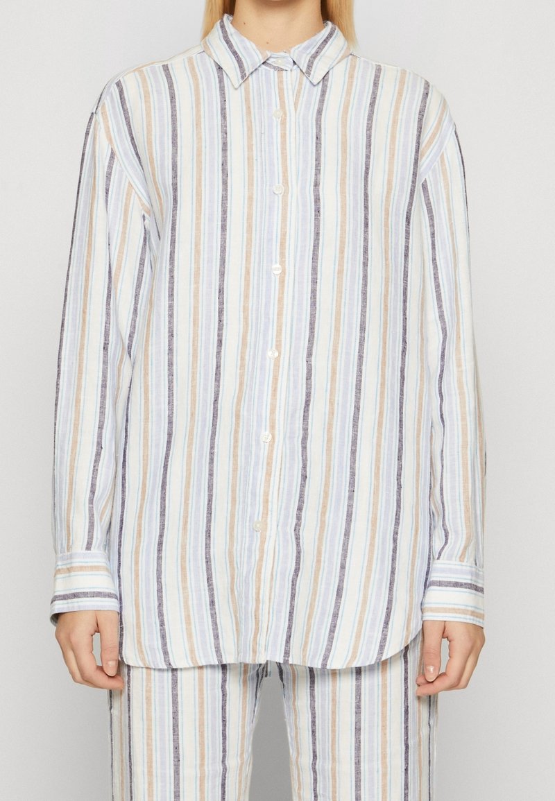 Personne portant une chemise blanche à manches longues boutonnée avec des rayures verticales beige, bleues et grises, assortie d’un pantalon rayé assorti.