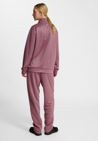 Roserød træningsdragt lavet af glat, syntetisk stof med høj krave, løstsiddende pasform og manchetter på både ærmer og bukser. Sorte sko vist.