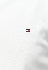 Tissu blanc texturé avec un petit logo rectangulaire comprenant des sections bleu marine, blanc et rouge brodées sur le côté droit.