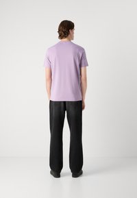 Camiseta de manga corta color lila con cuello redondo, combinada con jeans de pierna ancha negros, vista desde atrás sobre un fondo liso.