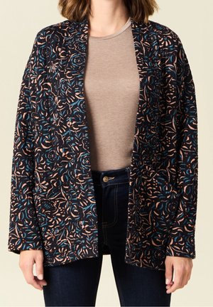 Donna che indossa una giacca trapuntata nera con motivi floreali blu e beige sopra una camicia beige e jeans blu scuro.