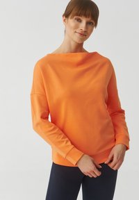 Maglietta arancione a maniche lunghe e vestibilità ampia con scollatura a barca e polsini a coste, realizzata in materiale morbido. Abbinata a pantaloni scuri.