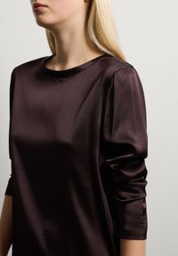 Mujer rubia vestida con una blusa de satén marrón oscuro de manga larga con mangas ligeramente recogidas, posando contra un fondo blanco puro.