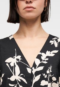 Femme aux cheveux courts et foncés portant un haut noir à col en V avec un grand imprimé floral blanc et de petits boutons noirs à l'avant.