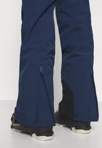 Marinblå vattentäta skidbyxor med blixtlås vid nedre benen och svarta detaljer, designade för utomhusbruk, kombinerade med svarta och lila kängor.