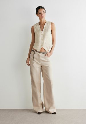 Femme portant un gilet beige sans manches boutonné et un pantalon beige à jambes larges avec une fine ceinture marron, debout contre un mur blanc uni.