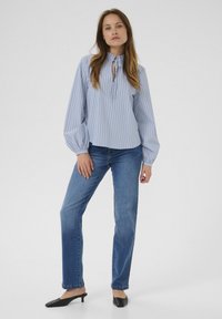 Camicia blusa azzurro chiaro con righe bianche verticali, scollatura a fiocco e maniche lunghe a sbuffo, abbinata a jeans blu a gamba dritta e mule nere.