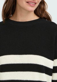 Svart og kremstripet genser med ribbestruktur, med bred utringning og lange ermer. Detaljer inkluderer kontrasterende horisontale striper.