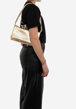 Metaliczna złota torebka o gładkiej fakturze, prostokątnym kształcie i jednym pasku na ramię, noszona z czarną koszulą i skórzanymi spodniami.