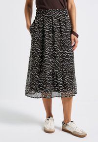 Femme portant une jupe midi à motifs noir et gris, des baskets blanches, un haut sombre et des bracelets sombres, se tenant avec une main dans la poche.
