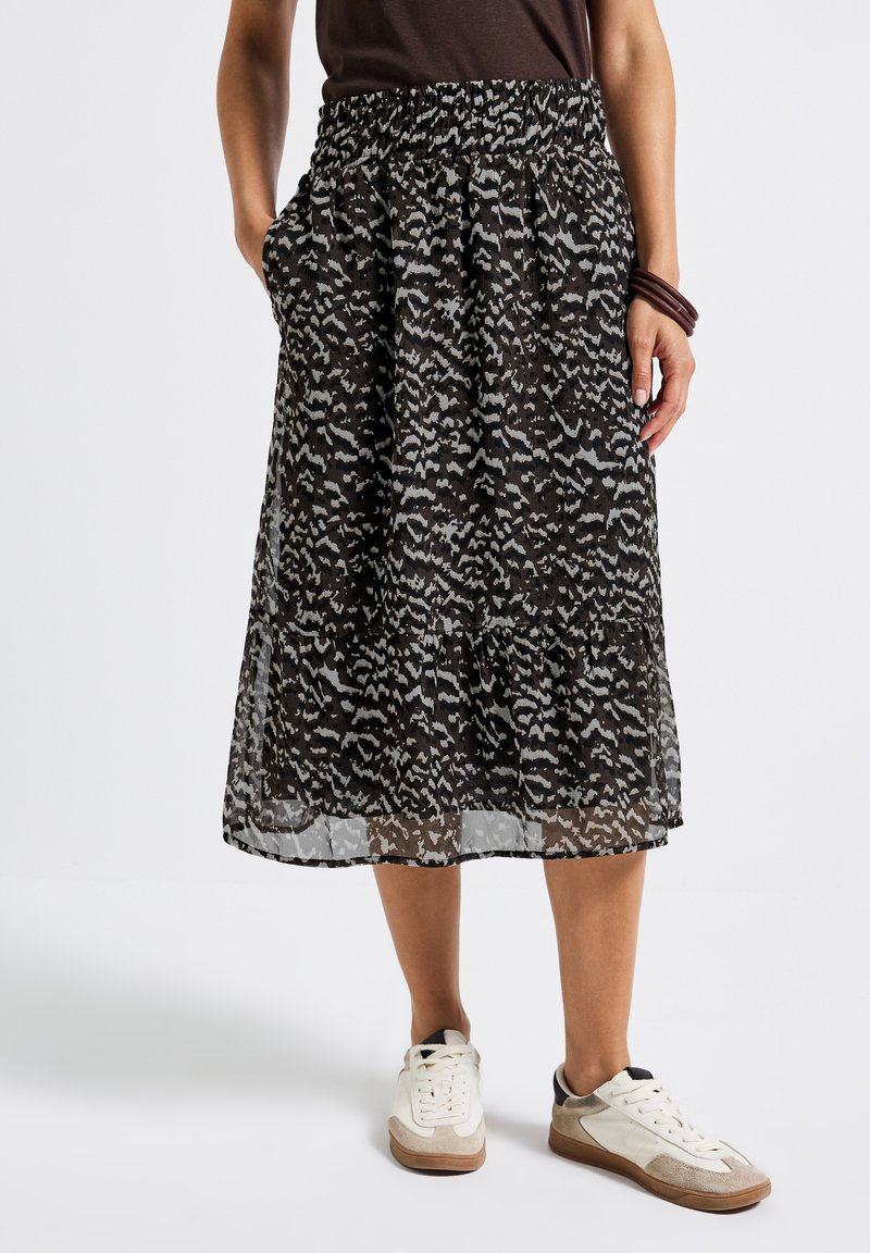 Femme portant une jupe midi à motifs noir et gris, des baskets blanches, un haut sombre et des bracelets sombres, se tenant avec une main dans la poche.
