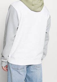 Huvtröja med vit kropp, grå ärmar och en mjuk olivgrön huva. Har en avslappnad passform och ribbad nederkant.