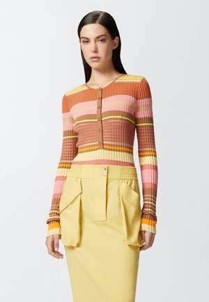 Cardigan multicolore à côtes avec col rond et boutons sur le devant, présentant des rayures marron, rose, jaune et orange. Associé à une jupe jaune.