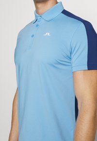 Ljusblå pikétröja med korta ärmar, kontrasterande marinblå detaljer, treknappskrage och liten vit logotyp på vänster bröst.